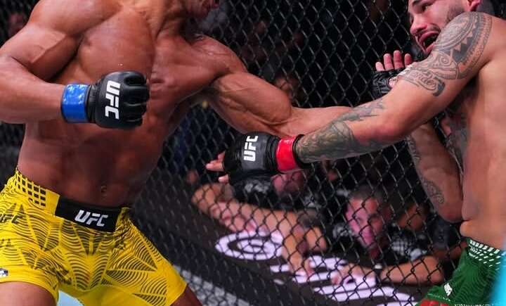 Two UFC fighters in a cage exchange punches; left fighter in yellow shorts lands a punch on the opponent in green shorts with red gloves at the Octagon edge.