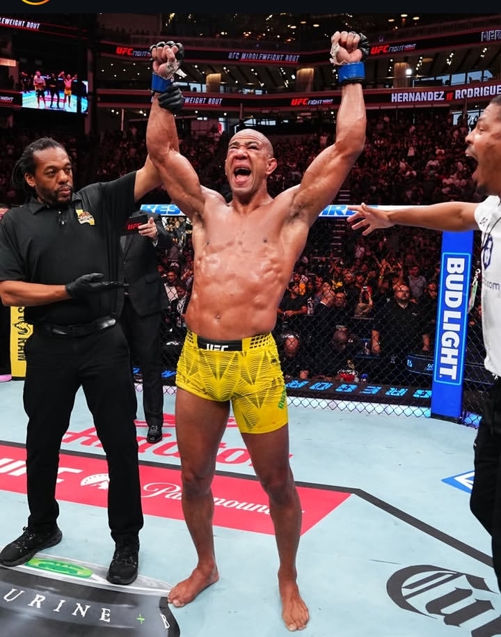 UFC fighter in yellow trunks raises his fists in victory inside the octagon, with a cheering crowd and an official nearby.