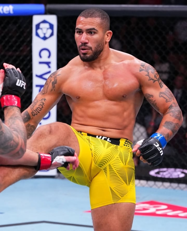 Male UFC fighter in bright yellow shorts and black gloves mid-action in the octagon, an opponent’s leg visible nearby.