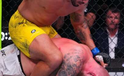 UFC-style ground fight: top fighter in yellow shorts delivering punches while pinning tattooed opponent on the mat inside the cage.