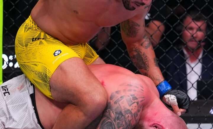 UFC-style ground fight: top fighter in yellow shorts delivering punches while pinning tattooed opponent on the mat inside the cage.