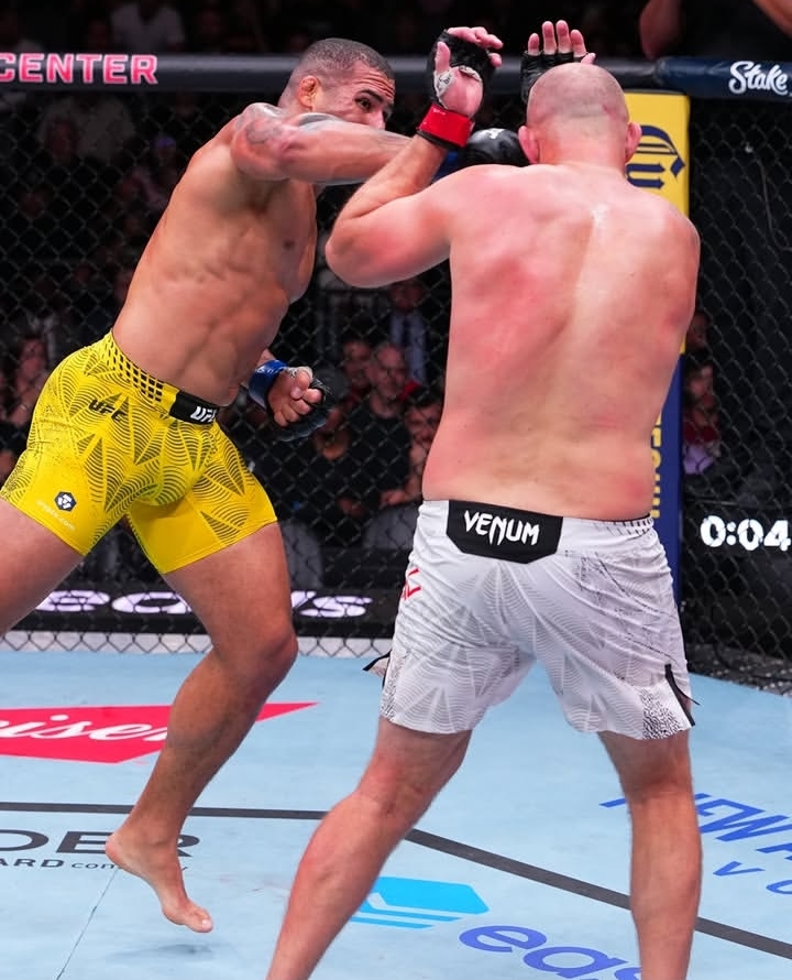 Two mixed martial artists exchange punches inside a fenced ring; fighter in yellow shorts lands a strike on the opponent in white Venom shorts while spectators watch.
