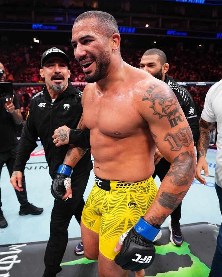 UFC fighter with yellow shorts and blue gloves smiling in the octagon after a fight, surrounded by teammates in the arena.