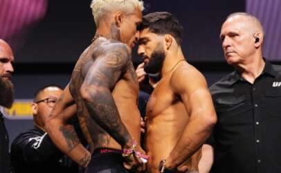 Two shirtless male fighters face off nose-to-nose on stage during a weigh-in, surrounded by officials and security. Tattoos visible on one fighter's arm and chest.