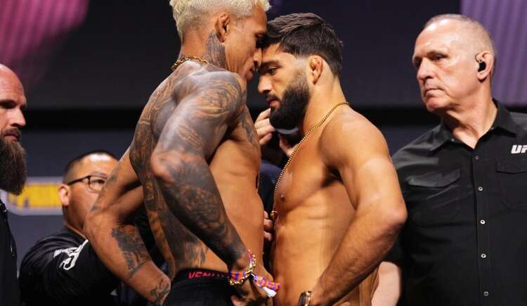 Two shirtless male fighters face off nose-to-nose on stage during a weigh-in, surrounded by officials and security. Tattoos visible on one fighter's arm and chest.