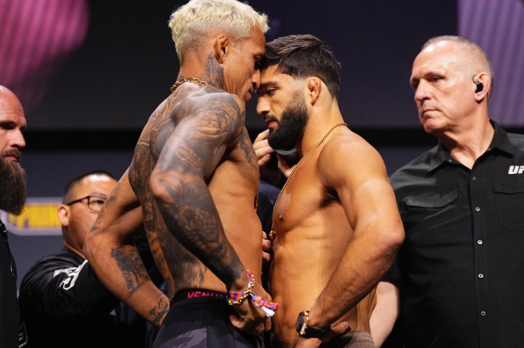 Two shirtless male fighters face off nose-to-nose on stage during a weigh-in, surrounded by officials and security. Tattoos visible on one fighter's arm and chest.