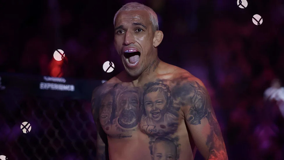 Shirtless male fighter with chest tattoos shouting inside a cage during a match in a dim arena.