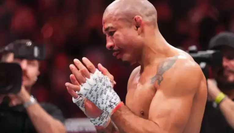 Bald male fighter wearing hand wraps applauding in an arena with cameras in the background.