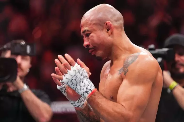 Bald male fighter wearing hand wraps applauding in an arena with cameras in the background.