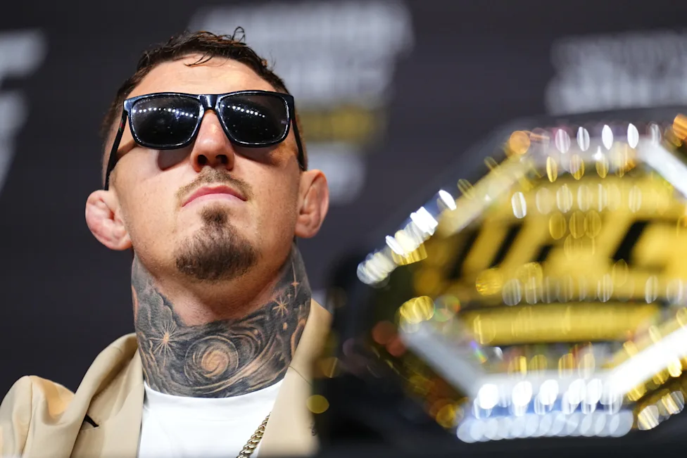 Man wearing dark sunglasses and a neck tattoo stands beside a gold trophy at a public event, looking upward with a confident expression.