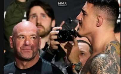 Bald UFC president Dana White smiles as a tattooed fighter aims a camera at him during a media event near photographers in the background.
