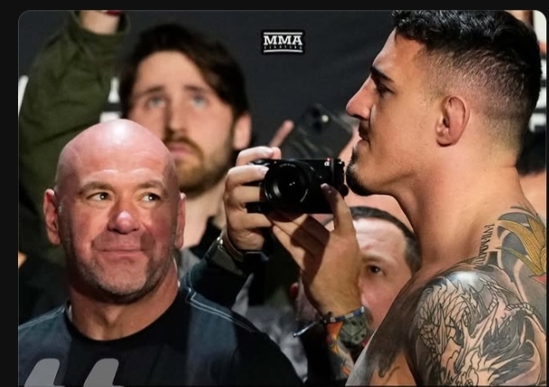 Bald UFC president Dana White smiles as a tattooed fighter aims a camera at him during a media event near photographers in the background.