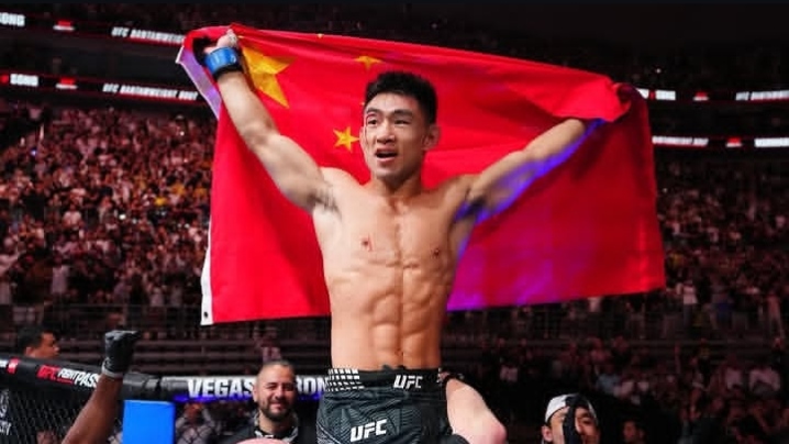 Male mixed martial artist in UFC shorts raises his arms in victory, draped in a Chinese flag behind him in the arena.