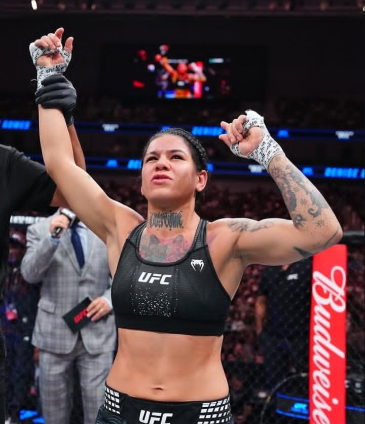Female UFC fighter in a black sports bra and shorts raises her arms in victory inside the octagon, gloves on and tattoos visible.