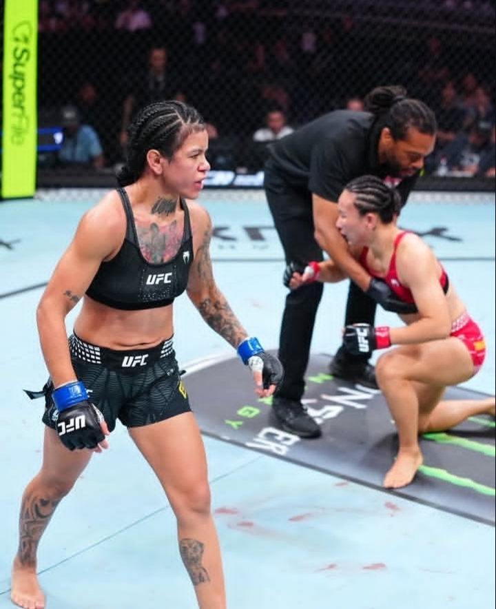UFC bout inside a cage: a female fighter in black gear walks toward the camera while her opponent in red kneels with a coach nearby.
