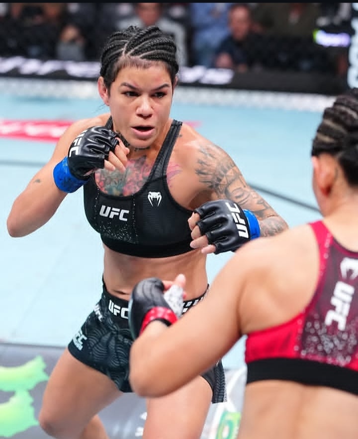 Female UFC fighter in a black sports bra and shorts throws a punch, tattoos on her left arm, cornrows, blue gloves inside the octagon.
