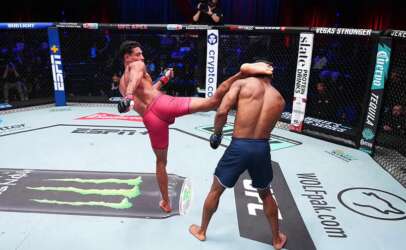Two male mixed martial artists spar in a fenced octagon; one delivers a kick to the other’s torso.