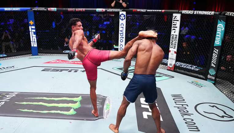 Two male mixed martial artists spar in a fenced octagon; one delivers a kick to the other’s torso.