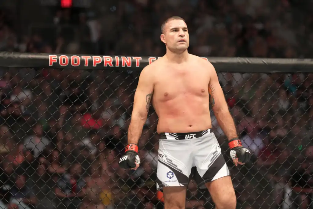 Male UFC fighter stands in the octagon shirtless with red gloves, looking ahead as spectators watch from the cage wall behind him.