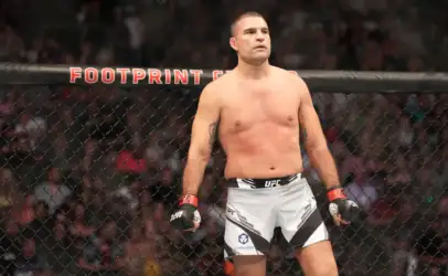 Male UFC fighter stands in the octagon shirtless with red gloves, looking ahead as spectators watch from the cage wall behind him.
