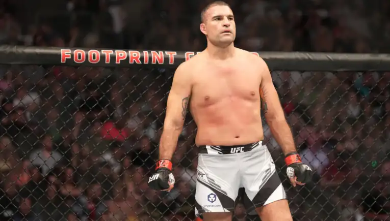 Male UFC fighter stands in the octagon shirtless with red gloves, looking ahead as spectators watch from the cage wall behind him.