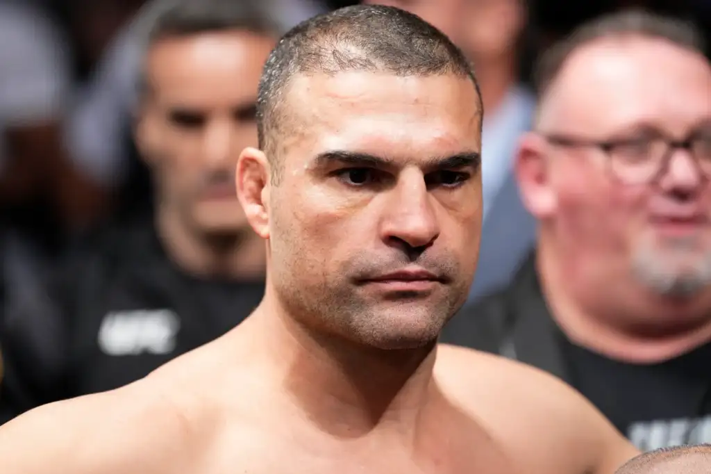 Close-up of a shirtless man with short hair and stubble, looking serious, with blurred people in the background.