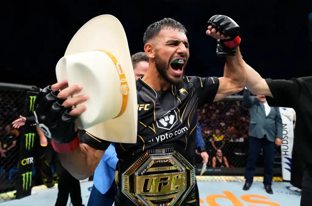 MMA fighter in black and gold gear celebrating inside the cage, fist raised, green mouthguard visible and championship belt around the waist.
