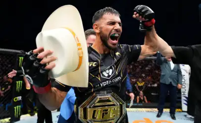 MMA fighter in black and gold gear celebrating inside the cage, fist raised, green mouthguard visible and championship belt around the waist.