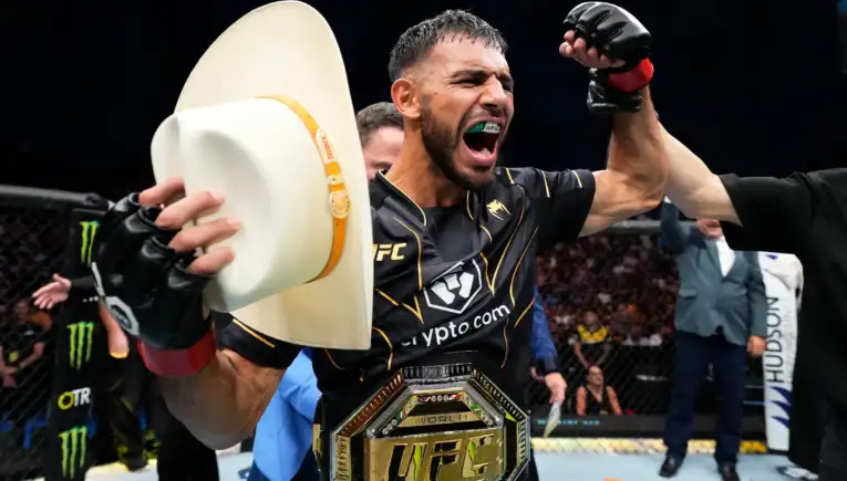 MMA fighter in black and gold gear celebrating inside the cage, fist raised, green mouthguard visible and championship belt around the waist.
