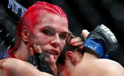 Female mixed martial artist with red braided hair, sweaty face, and gloves resting near her mouth in the octagon, looking intense and exhausted after a fight.