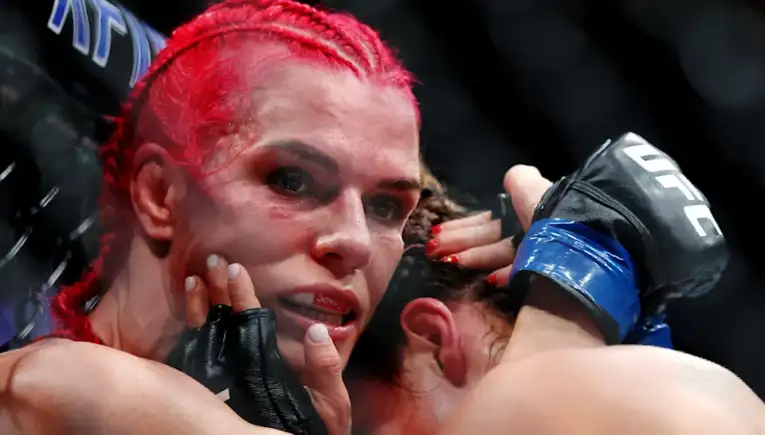 Female mixed martial artist with red braided hair, sweaty face, and gloves resting near her mouth in the octagon, looking intense and exhausted after a fight.