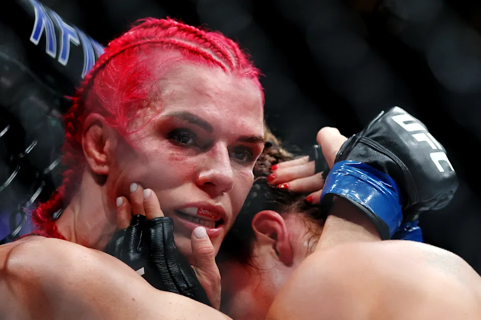 Female mixed martial artist with red braided hair, sweaty face, and gloves resting near her mouth in the octagon, looking intense and exhausted after a fight.