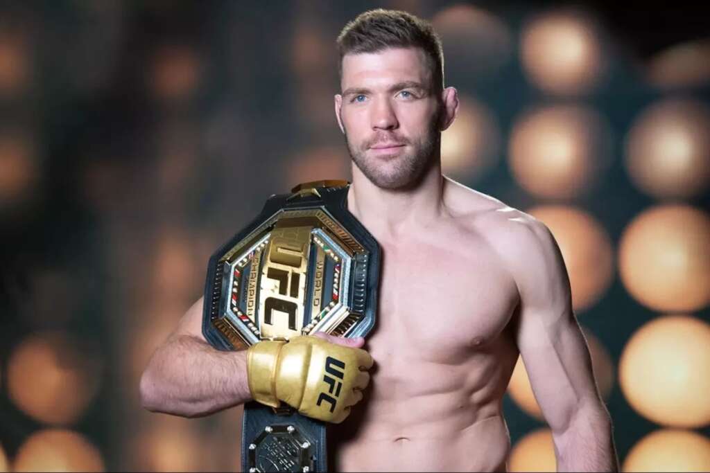 Male mixed martial artist posing shirtless with a UFC championship belt draped over his shoulder in a blurred arena backdrop.