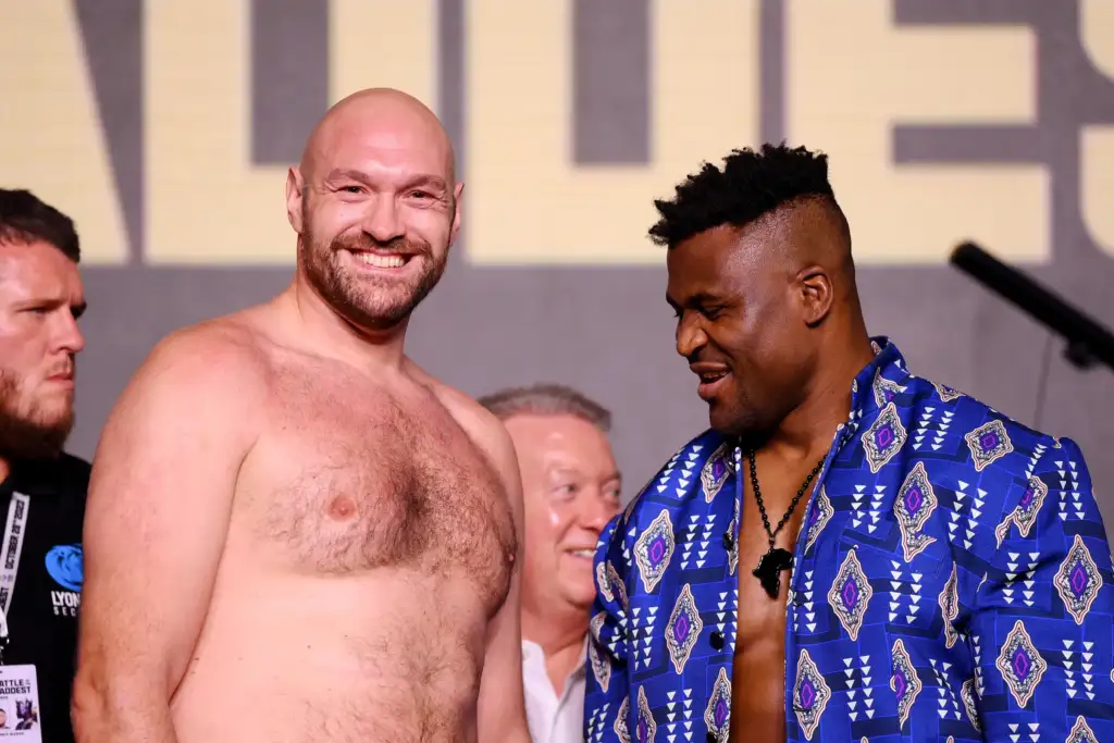 Smiling bald, shirtless man stands next to a man in a blue patterned shirt at a stage event, with spectators in the background.