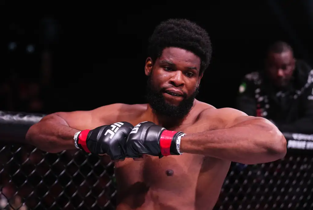 Male mixed martial artist with a beard rests his arms on the cage, shirtless, in a dimly lit octagon.