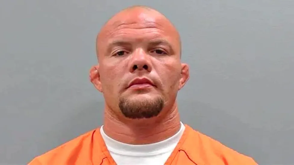 Mugshot of a bald man with a goatee wearing an orange jumpsuit against a gray backdrop