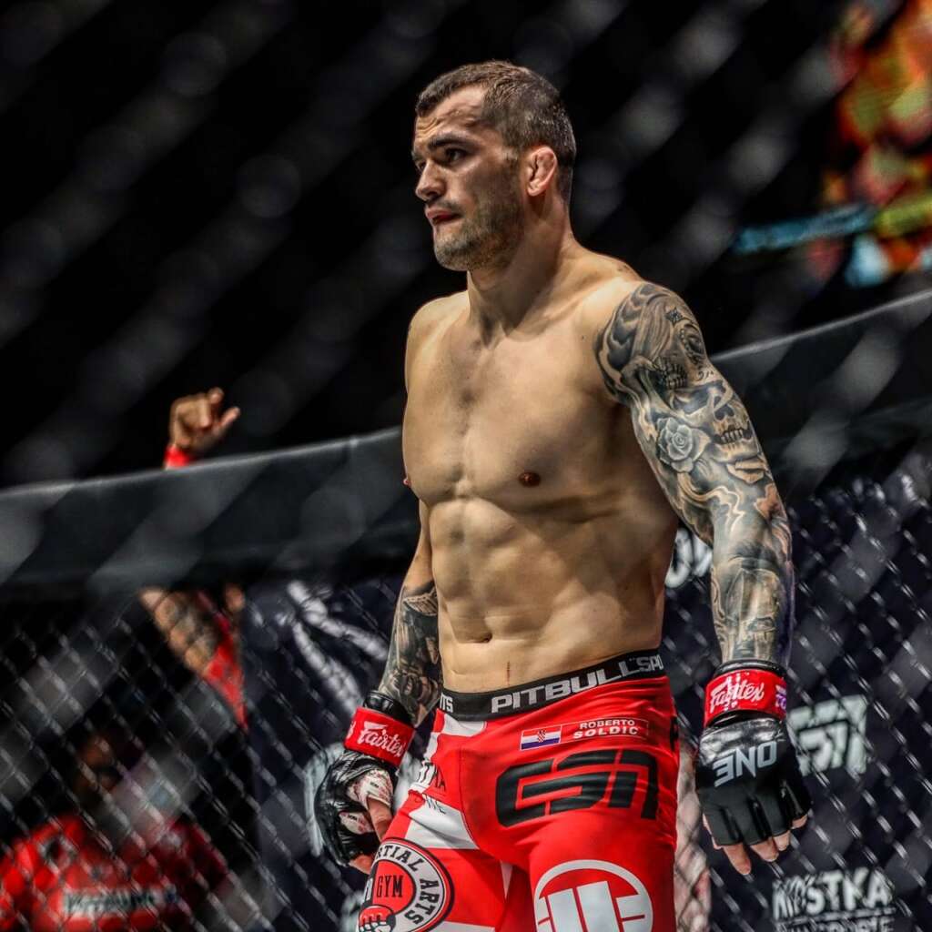 MMA fighter standing in the cage in red shorts and black gloves, muscular with a tattoo sleeve on his left arm.