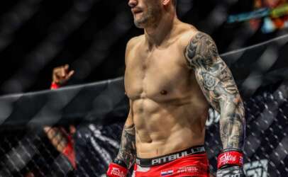 MMA fighter standing in the cage in red shorts and black gloves, muscular with a tattoo sleeve on his left arm.