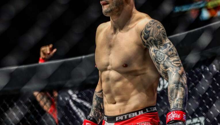 MMA fighter standing in the cage in red shorts and black gloves, muscular with a tattoo sleeve on his left arm.