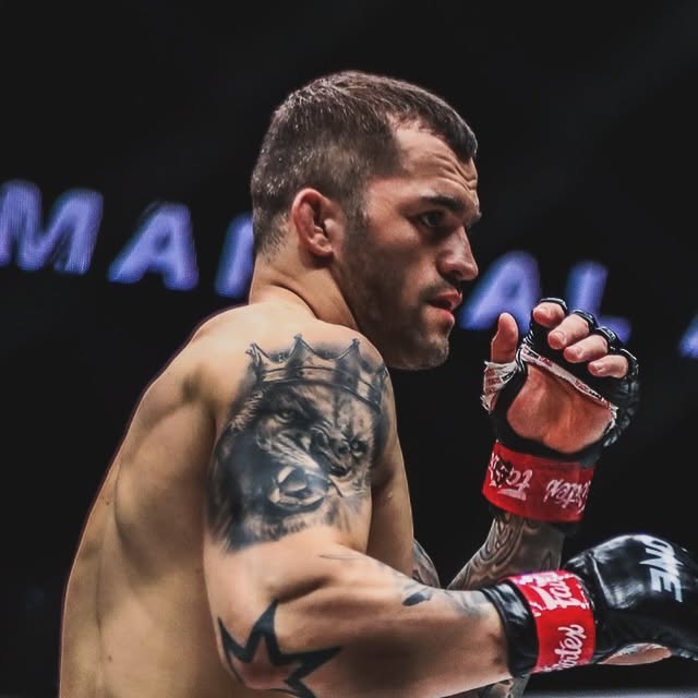 Male mixed martial artist in red gloves turns toward the camera, showing a tiger tattoo on his upper arm before a fight relief shot.