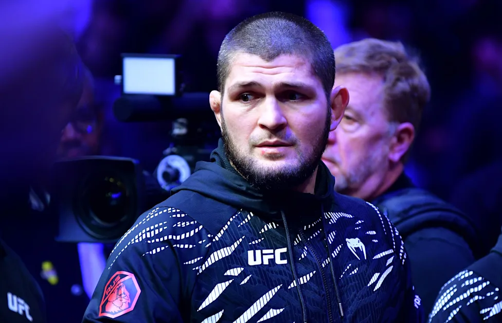 UFC fighter in a patterned jacket pauses backstage as a camera crew works under blue lighting.