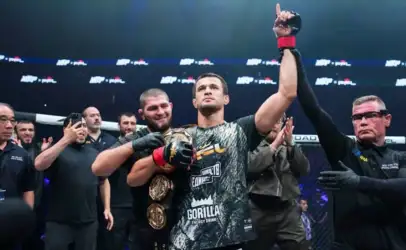 Male mixed martial artist raises his arm in victory as the referee holds his wrist inside the ring, crowd applauding behind them.