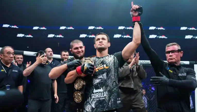 Male mixed martial artist raises his arm in victory as the referee holds his wrist inside the ring, crowd applauding behind them.