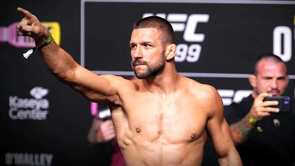 Shirtless male UFC fighter on a weigh-in stage points toward the crowd with a focused expression, behind him a UFC backdrop and spectators.