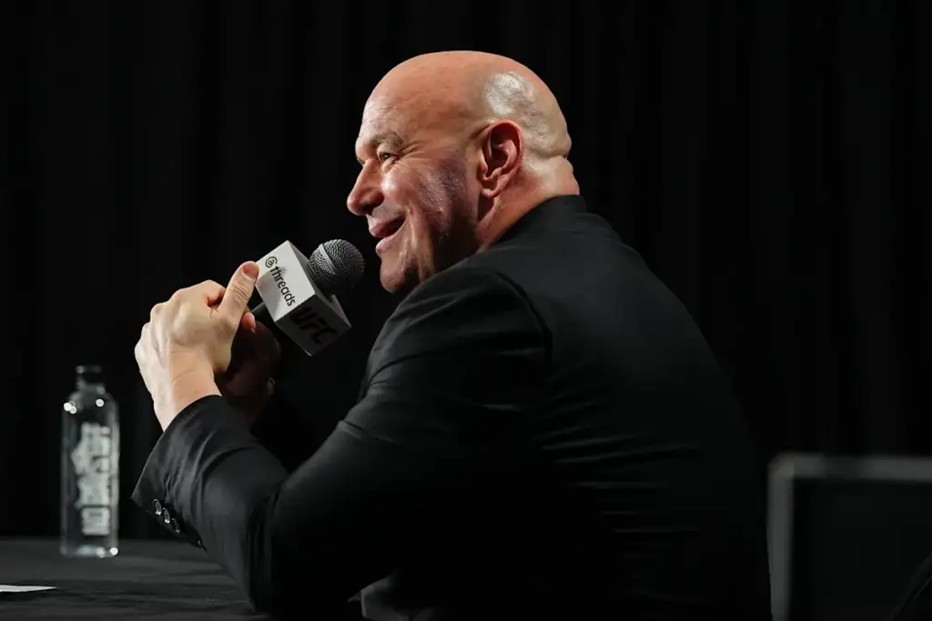 Bald man in a dark suit speaks into a microphone labeled UFC at a press event, with a water bottle on the table nearby.