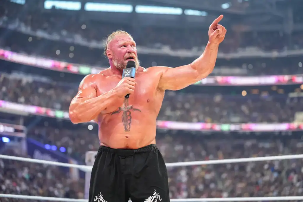 Wrestler with a chest tattoo speaks into a microphone in the ring, pointing upward to the crowd.