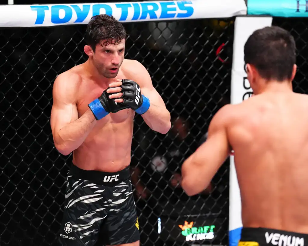 Two male UFC fighters in a cage, facing off; the foreground fighter wears blue hand wraps and patterned black shorts as he guards with his hands up.