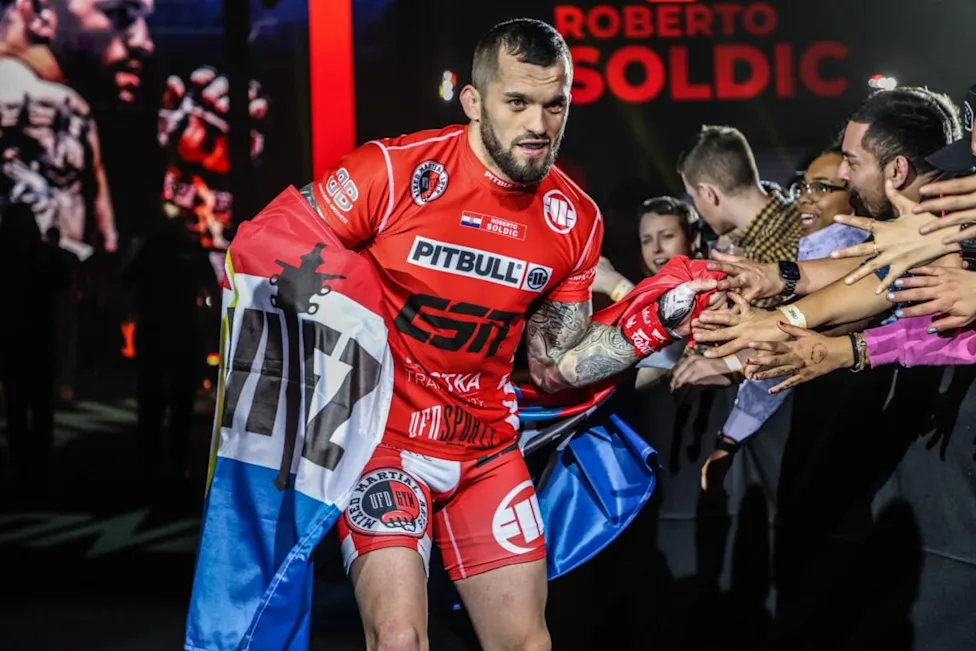 MMA fighter in red gear greets cheering fans, shaking hands as he enters the arena, with a blue flag draped over his shoulders and ‘ROBERTO SOLDIC’ sign in the background.