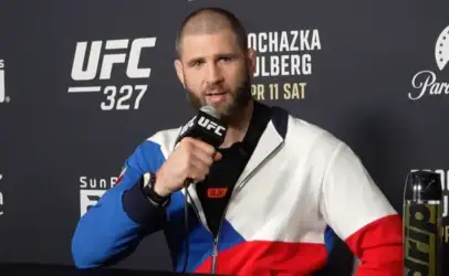 MMA fighter speaks into a microphone at a UFC press event, wearing a color-block jacket against a sponsorship backdrop.