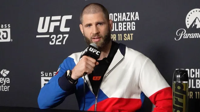 MMA fighter speaks into a microphone at a UFC press event, wearing a color-block jacket against a sponsorship backdrop.